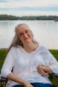Picture of Amanda. She is a white woman wearing a white shirt and glasses. Her long hair is pulled back from her face, and she is smiling at the camera with her head tilted to the left. In the background there is green grass with a calm lake just behind the grassy area. 