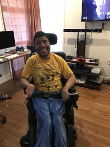 Image of Brent sitting in his wheelchair. He is an African American/Black male with short black hair. He is wearing a yellow long sleeve shirt and jeans. Behind and to his left is a computer desk with his monitor visible. Behind him is an entertainment stand with cords and game controllers visible. Mounted on the wall is a television. 