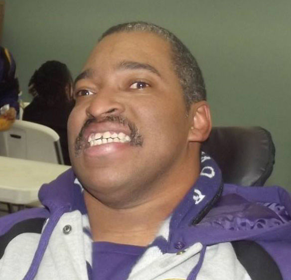 Photo of Jamal. He is a Black man with a mustache. He is sitting in his wheelchair and wearing a purple, black and white jacket. He is smiling at the camera.