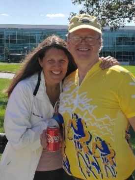 Image of Lisa Derx standing outside with a white man wearing a yellow shirt, baseball cap and sunglasses. Lisa has her arm around the man, her long brown hair is blowing in the wind, and she is wearing a white jacket. Lisa is smiling broadly at the camera.