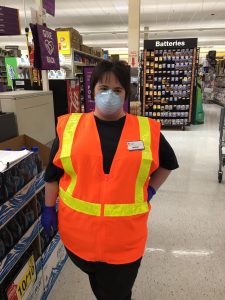 Meghan is standing in a grocery store isle. She has short brown hair with bangs and is wearing an N-95 mask. She is also wearing a black t-shirt with an orange reflective vest over top of it. She has one gloved hand in her pocket and the other is by her side. 