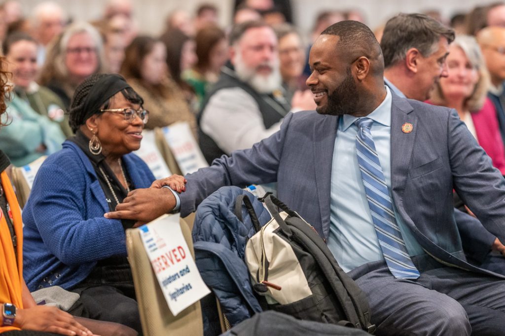 Senator Cory McCray turns around to chat with a woman sitting behind him. They are smiling at each other. Photo by Jennifer Bishop.