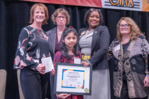 A young girl stands on stage after winning an award for the Together We're Better artwork contest. She is joined on stage by representatives from MSDE, MDOD, the DD Council, and The Arc Maryland.